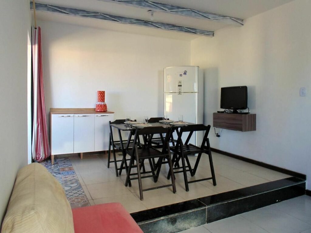Uma cozinha e espaço para refeição no FAROL BEACH Apartamentos & Suítes. No canto inferior esquerdo da foto é possível ver parte de um sofá. Mais para a frente, vemos uma mesa preta com quatro cadeiras, e ao seu lado direito há uma prateleira na parede com TV. Uma geladeira e um armário estão encostados na parede do fundo.