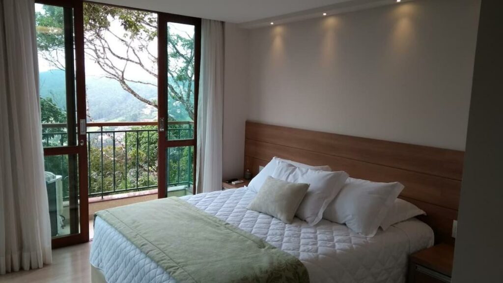 Quarto do Flat Vista Azul Hotel com uma cama de casal no meio e ao seu lado esquerdo uma varanda coom vista para a natureza.
