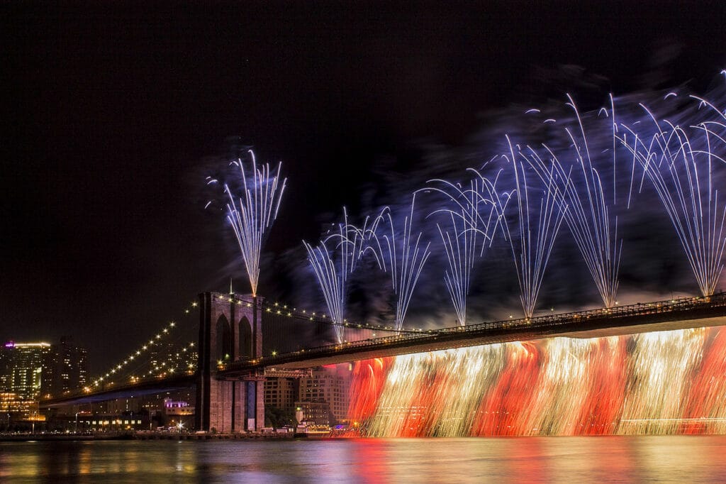Uma ampla ponte, a Brooklyn Bridge, inteira iluminada de noite e com fogos de artíficio sendo lançados dela e iluminando o céu escuro