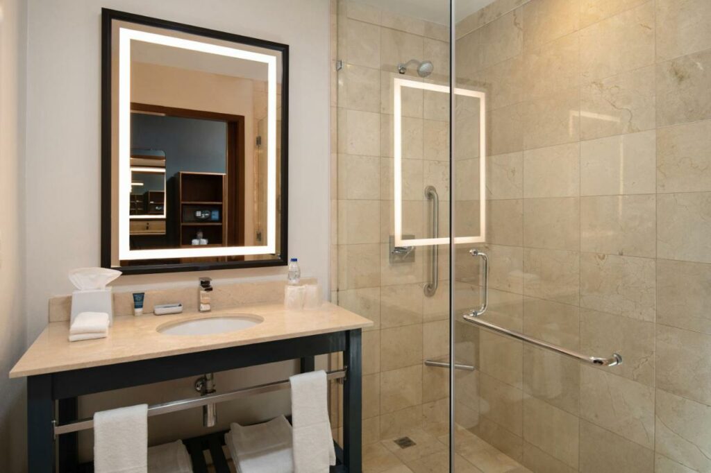 Banheiro do Four Points by Sheraton com um box de vidro que tem barras de apoio na área do chuveiro. Uma pia com amenidades de banho, toalhas e espelho fica do lado esquerdo do box.