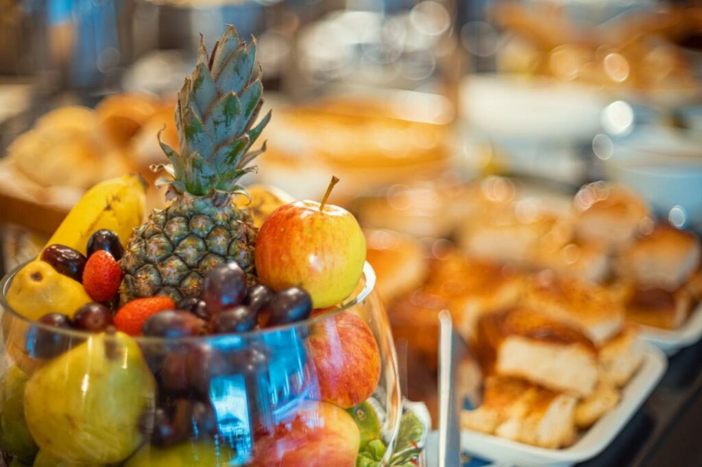 Foto do café da manhã do Grande Hotel da Barra. Em foco, há uma tigela de vidro completa com diversas frutas como abacaxi, maçãs, uvas, morango e peras. A imagem no fundo está desfocada, mas percebemos que há outras massas, como alguns pães.