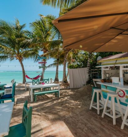 Estrutura do clube de praia do Green Coast Beach Hotel, um dos melhores hotéis em Punta Cana. Um bar de madeira colorido tem banquinhos em frente e fica no canto direito da imagem, com mesas e cadeiras na areia logo em frente. Ao fundo é possível ver o mar cristalino e palmeiras com redes penduradas.