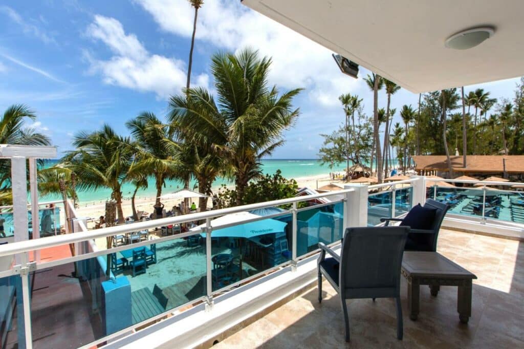 Vista do terraço do Green Coast Beach Hotel, que é coberto e tem mesinha e cadeiras. Ao fundo está uma praia de areia cheia de árvores, mesas e turistas, e mar cristalino.