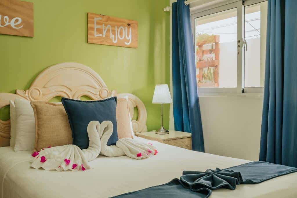 Cama de um quarto do Green Coast Beach Hotel, um dos hotéis perto do aeroporto de Punta Cana, que tem mesa de cabeceira com abajur do lado direito e toalhas dobradas em forma de cisne encostadas nos travesseiros. Há uma janela com cortinas na parede do lado direito, e dois quadrinhos na parede acima da cama.