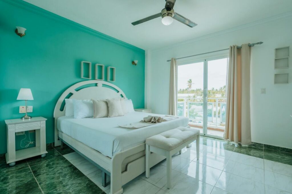 Quarto do Green Coast Hotel, um dos hotéis em Punta Cana, decorado em tons de branco e verde-água. Uma cama de casal fica encostada na parede do lado esquerdo e tem mesinhas com abajures dos dois lados e um banquinho aos pés. Há um ventilador no teto, e uma janela de vidro do lado direito da cama tem vista para fora do hotel.
