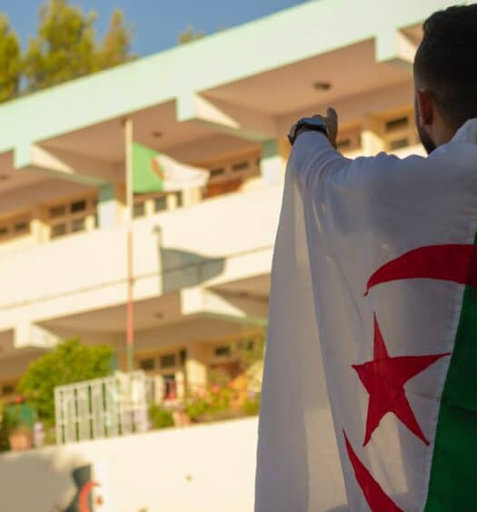 Homem do lado direito com a bandeira da Argélia nas costas e ao fundo uma bandeira. Representa seguro viagem Argélia.