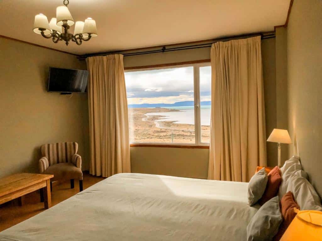 Quarto da Hosteria La Estepa. Uma cama de casal e um abajur do lado direito, de frente uma mesa, uma poltrona e uma televisão. No fundo, uma janela com cortina e vista para o Lago Argentino.
