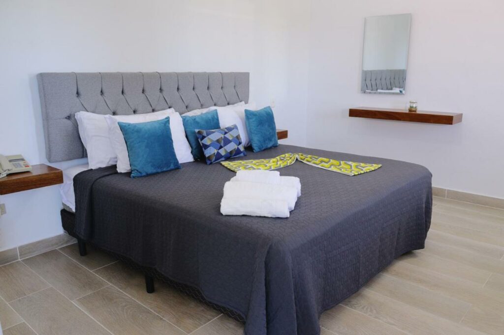 Cama em quarto do Hotel Capriccio Mare y Restaurante, um dos hotéis em Punta Cana. Há apoios de cabeceira dos dois lados da cama de casal, que é repleta de almofadas e tem toalhas dobradas aos pés. Um pequeno espelho fica ao lado direito.