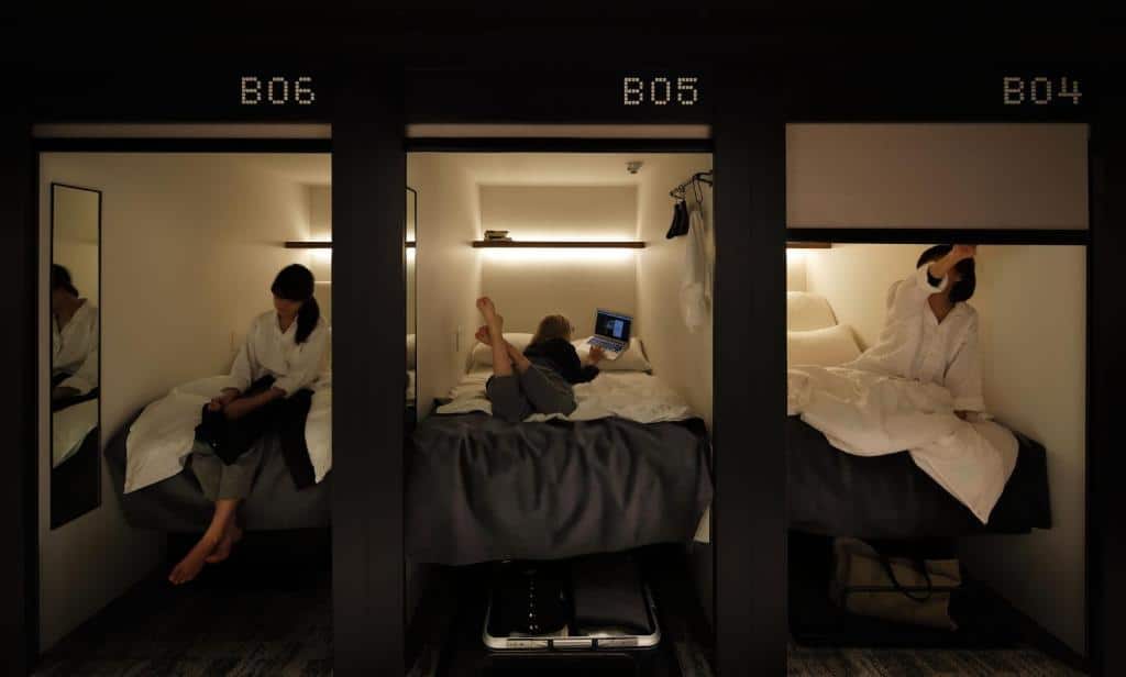 Foto do hotel The Millennials Shibuya. Na imagem vemos três cápsulas do hotel, todos com suas respectivas camas. Há uma mulher em cada um deles. Na primeira cápsula há uma mulher está sentada na cama enquanto dobra suas roupas, na segunda há uma mulher deitada mexendo em seu computador, e na terceira tem uma mulher que está abaixando a cortina, preparando-se para dormir.