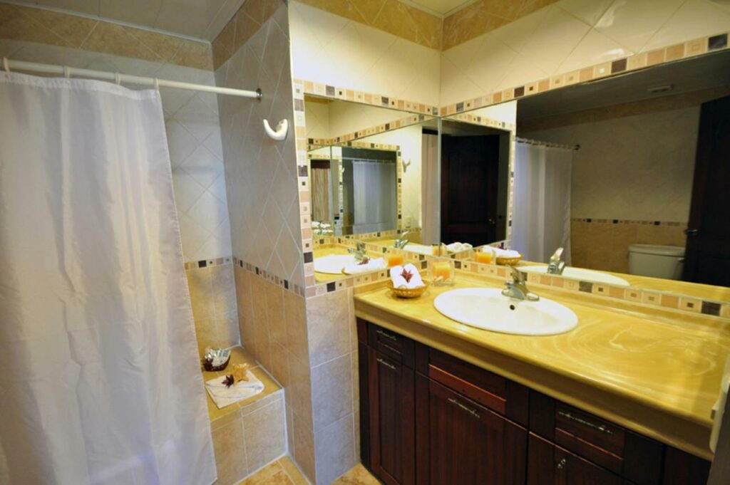 Banheiro do Hotel & Casino Flamboyan. Do lado direito da imagem, uma pia com amenidades de banho e espelho reflete a imagem da privada em frente. Do lado esquerdo, uma banheira com chuveiro tem uma cortina fechada.
