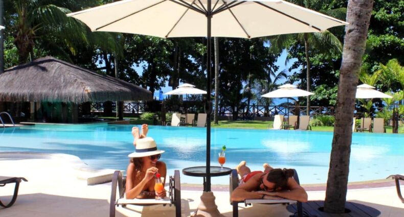 Uma mulher e um homem deitados em uma espreguiçadeira no Iara Beach Hotel Boutique enquanto bebem drinks. Sobre eles, há um guarda-sol, e, ao fundo, vemos a piscina e outros guarda-sóis ao redor dela, para representar hotéis para o Carnaval em Salvador