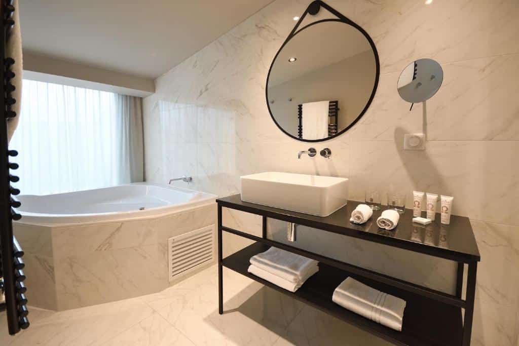 Banheiro do Hôtel La Maison Bord'eaux. Ao fundo há uma banheira e logo ao lado há uma pia com toalhas e produtos de banho. Acima dela há dois espelhos de formato redondo, um grande e um menor.