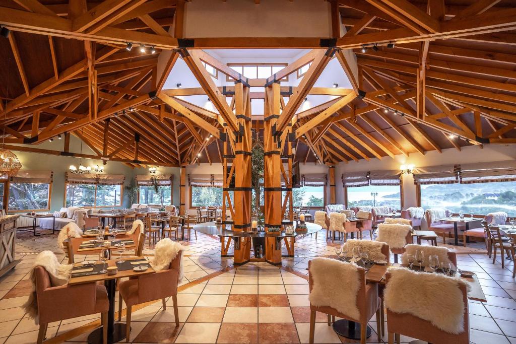 Restaurante do Hotel Los Ñires. Cadeiras com encosto e coberta de pelo, no centro uma ilha para apoio da comida e uma arquitetura de madeira. O restaurante com paredes de vidro e vista para as montanhas.