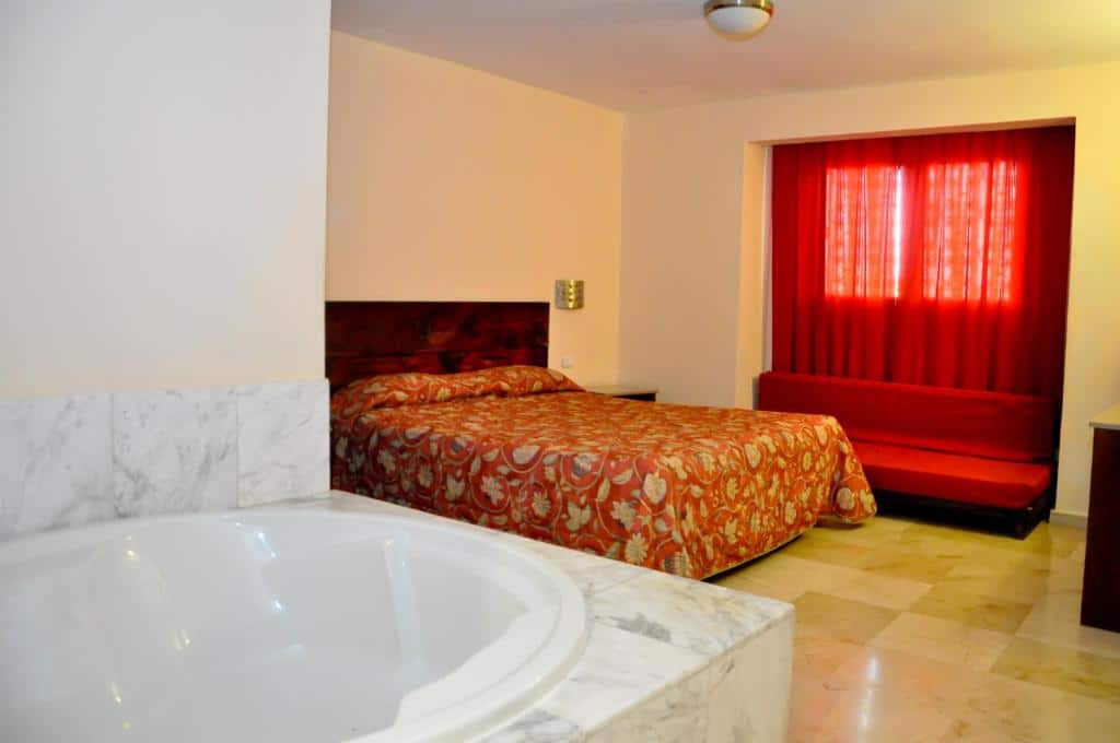Quarto do Hotel Marimba Punta Cana, um dos hotéis em Punta Cana. Em primeiro plano fica uma banheira de hidromassagem, e ao fundo uma cama com mesinhas de cabeceira dos dois lados está encostada na parede do lado direito. Uma janela está coberta por cortinas vermelhas do lado direito da cama.