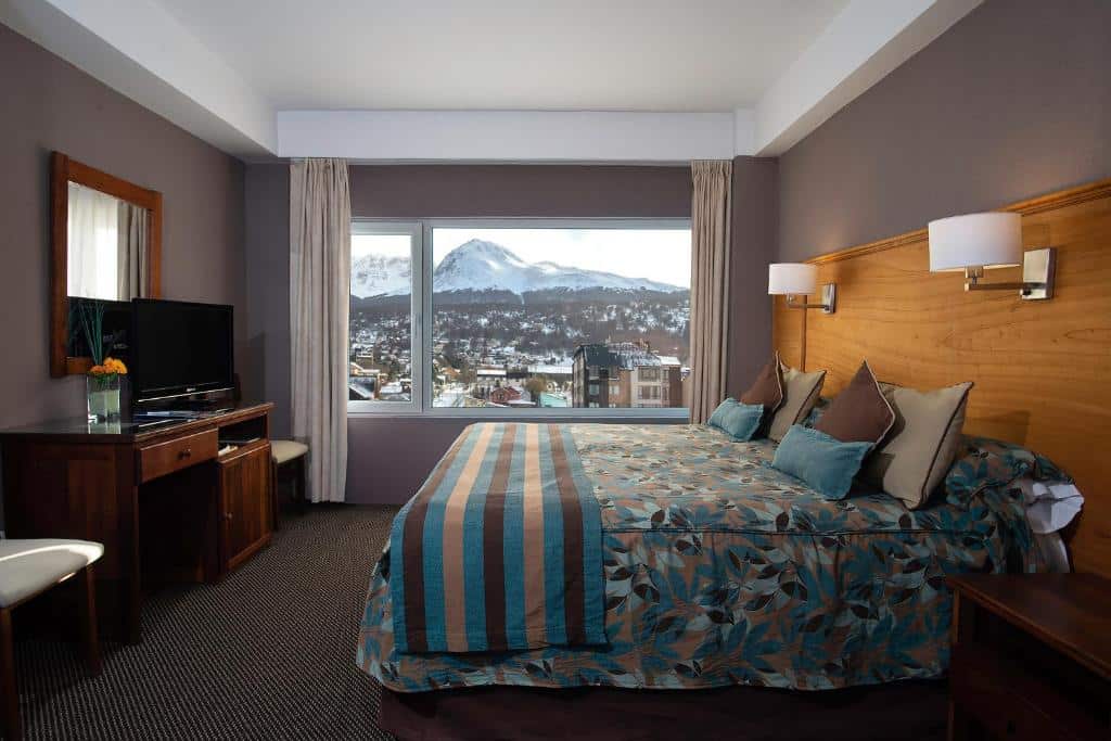 Quarto do Hotel Tierra del Fuego. Uma cama de casal do lado direito com um abajur de cada lado e uma cômoda. De frente uma televisão com uma raque, um espelho e duas cadeiras. No fundo, a janela do quarto com vista para a cidade e neve. Foto para ilustrar post sobre hotéis em Ushuaia.