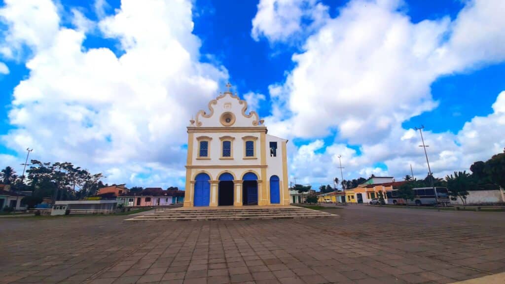 Igreja do Senhor do Bonfim, em Marechal Deodoro. A igreja fica em uma praça com área extensa à frente, e tem uma arquitetura histórica, com as bordas amarelas e portas azuis. Há casas históricas nas ruas ao lado