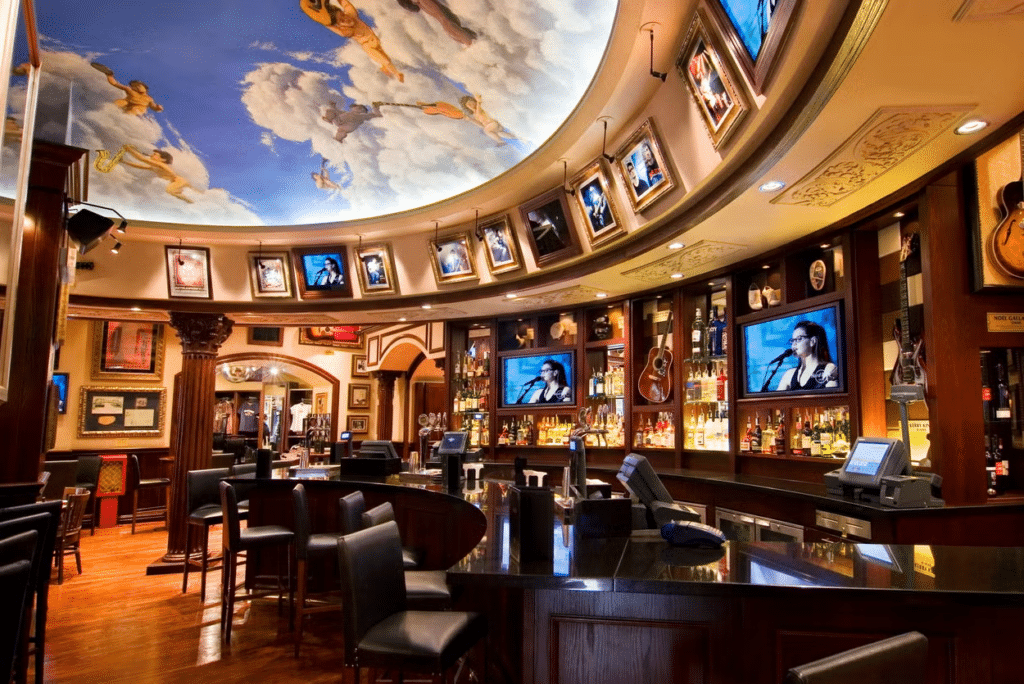 vista do interior do Hard Rock Cafe em Roma , com balcão e poltronas estofadas, televisões com música rolando, diversas bebidas e teto temático que imita um afresco de anjinhos no céu com nuvens