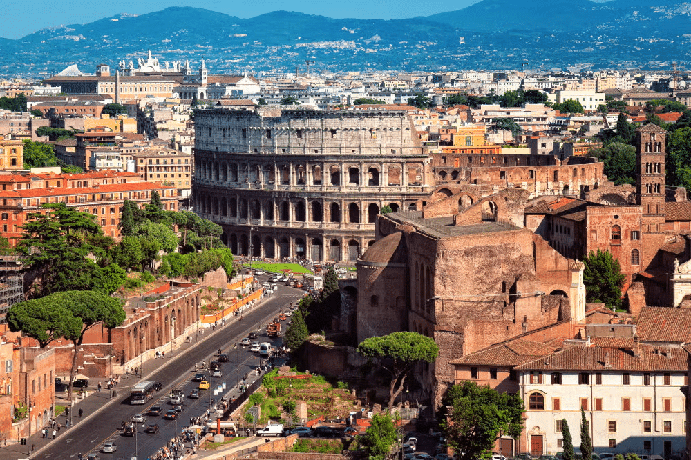 vista do exterior do Coliseu, em Roma com uma avenida grande com muitos carros chegando até a atração, há casas históricas e uma bela paisagem atrás