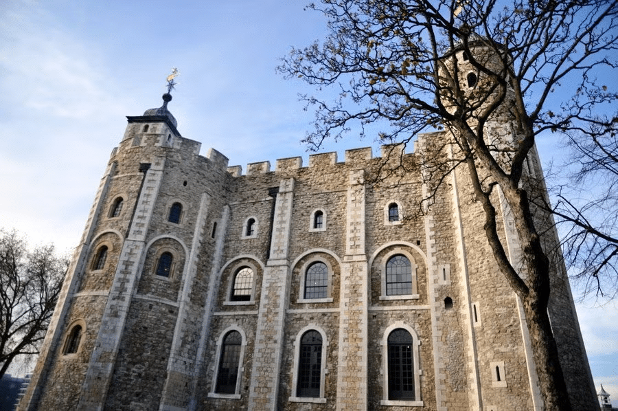 Vista de baixo para cima da Torre de Londres, um monumento imponente em pedra, com janelas em arcos, que lembra um castelo, há árvores ao redor