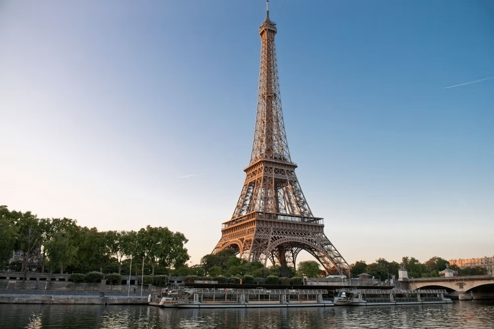 Vista da Torre Eiffel às margens do rio Sena a partir do barco, há uma ponte e várias árvores ao redor.