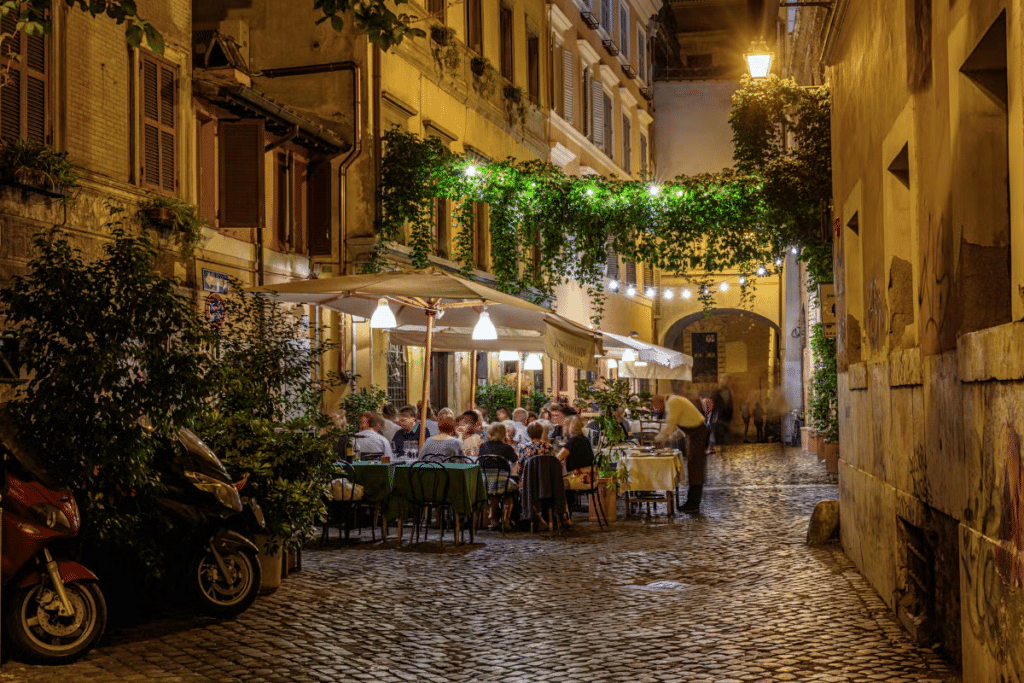 Rua com calçada de pedras com edifícios com várias janelinhas de madeira, há varais de plantas e mesinhas embaixo de uma tendinha no bairro do Trastevere, uma das opções de o que fazer em Roma