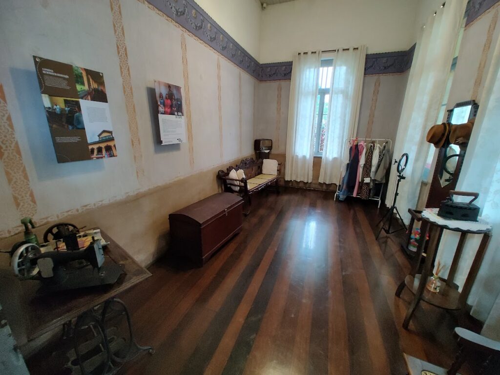 sala interior com piso de madeira e móveis antigas do século 19 e 20, incluindo uma mesa de costura, um ferro de passar roupa, um baú, um rádio e algumas roupas.