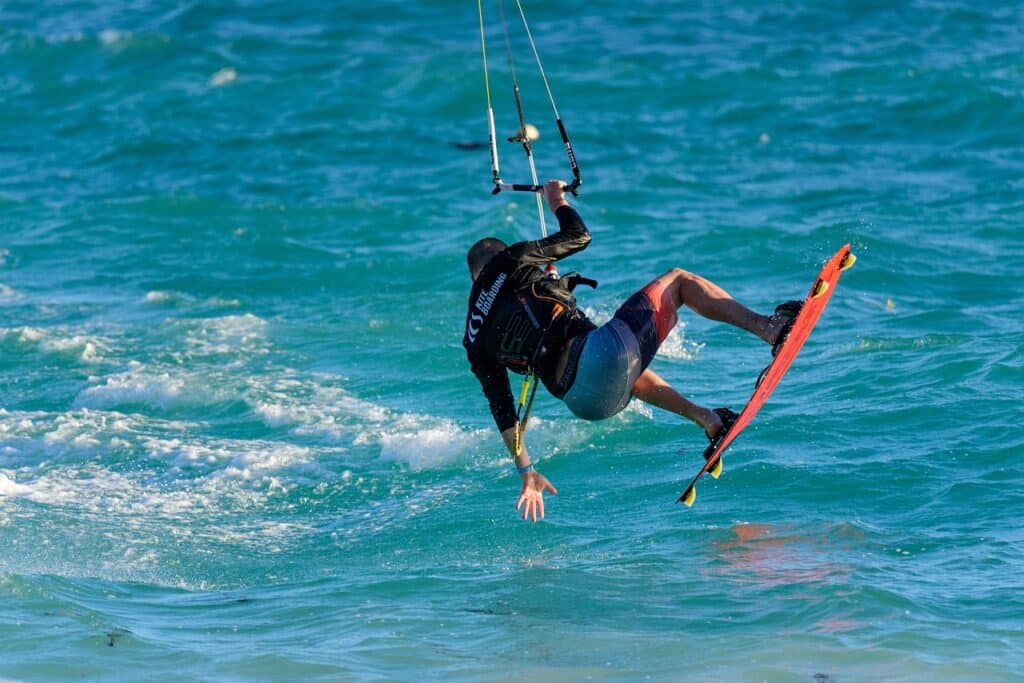 Homem de costas em cima de uma prancha e segurando um apoio aéreo enquanto pula nas ondas do mar logo abaixo. - Foto: MustangJoe via Pixabay