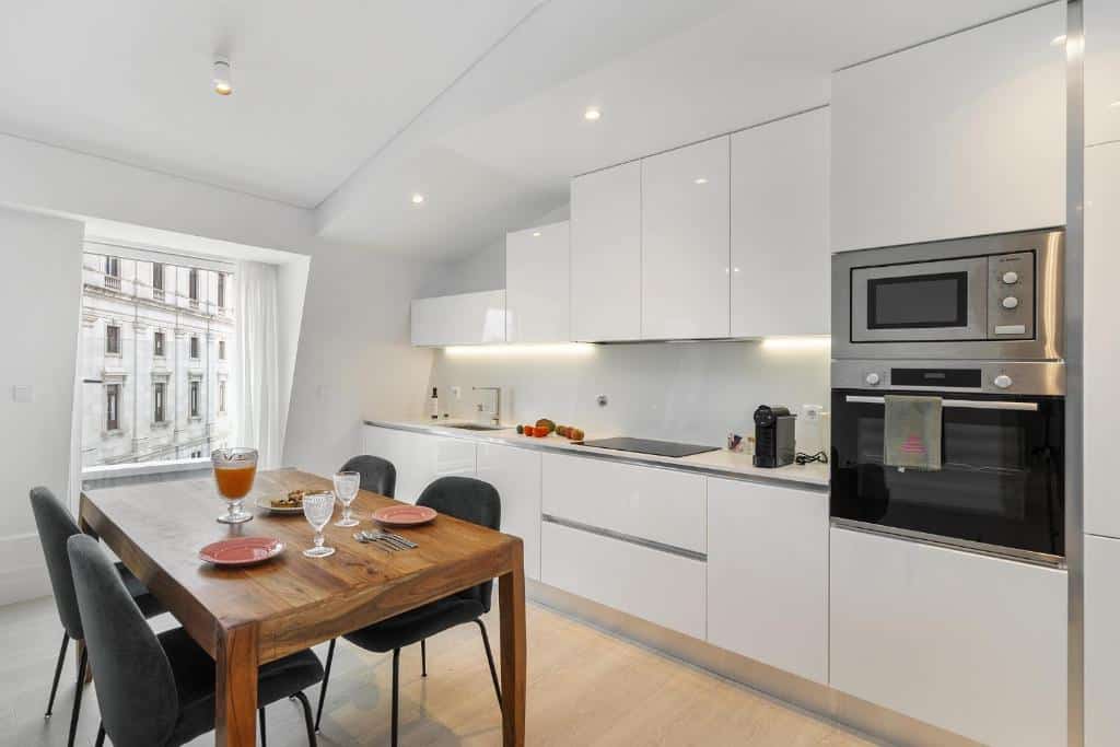 Cozinha de um dos apartamentos do Les Deux Mariettes São Bento com uma cozinha com armários brancos, uma torre com forno e micro-ondas, além de uma janela do lado esquerdo e uma mesa de madeira com quatro cadeiras estofadas