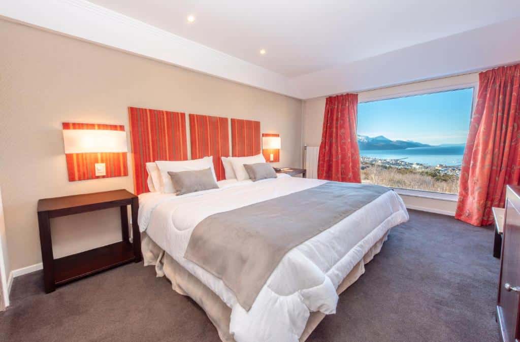 Quarto do Los Acebos Ushuaia Hotel. Uma cama de casal do lado esquerdo, com uma mesa de apoio de cada lado, no fundo, uma parede de vidro com cortinas e vista para o lago. Foto para ilustrar post sobre hotéis em Ushuaia.