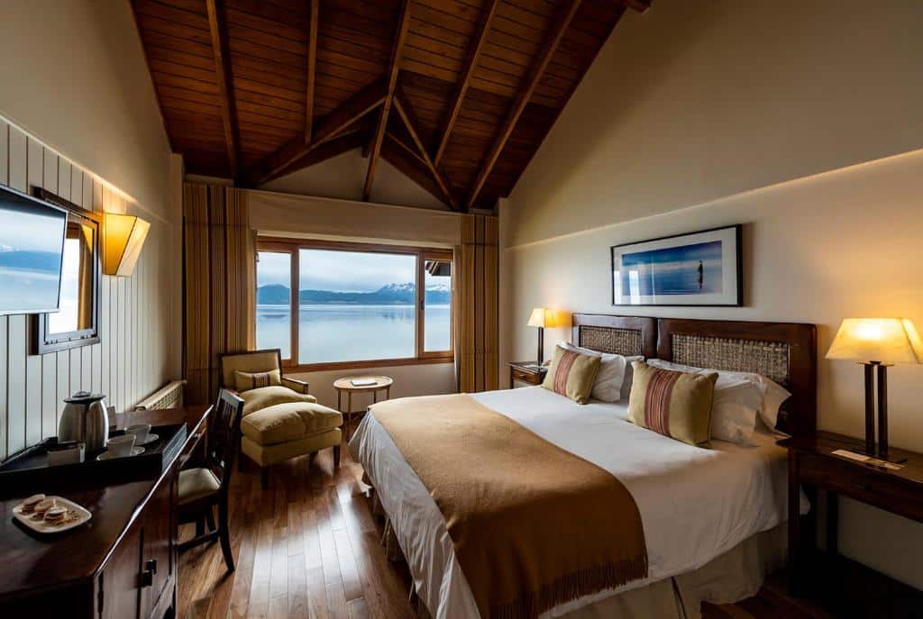 Quarto do Los Cauquenes Resort + Spa + Experiences em Ushuaia, com uma cama de casal do lado direito, com um abajur de cada lado, de frente para a cama uma televisão, uma mesa de trabalho com xícaras de café e um prato de doce. No fundo do quarto, uma poltrona, uma mesa e a janela do quarto.