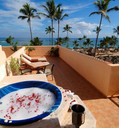 Sacada de um quarto do Majestic Colonial Punta Cana, um dos hotéis românticos em Punta Cana. Do lado esquerdo há uma banheira de hidromassagem com pétalas de rosa e um balde com garrafa de champanhe e taças ao lado. Há espreguiçadeiras ao fundo, e o lugar tem vista para a praia e palmeiras altas.