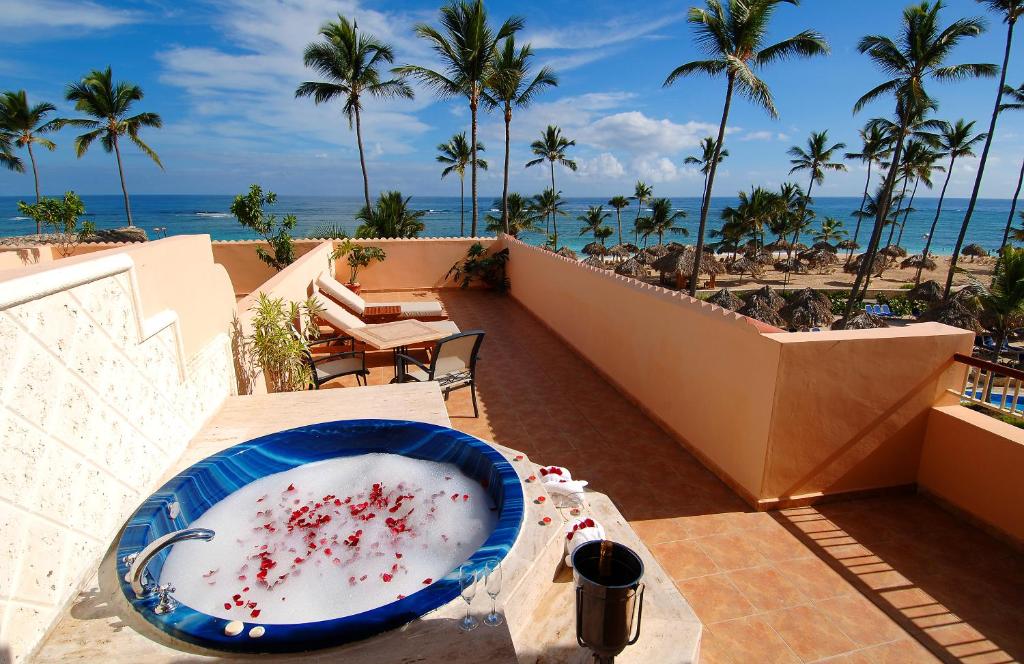 Sacada de um quarto do Majestic Colonial Punta Cana, um dos hotéis românticos em Punta Cana. Do lado esquerdo há uma banheira de hidromassagem com pétalas de rosa e um balde com garrafa de champanhe e taças ao lado. Há espreguiçadeiras ao fundo, e o lugar tem vista para a praia e palmeiras altas.
