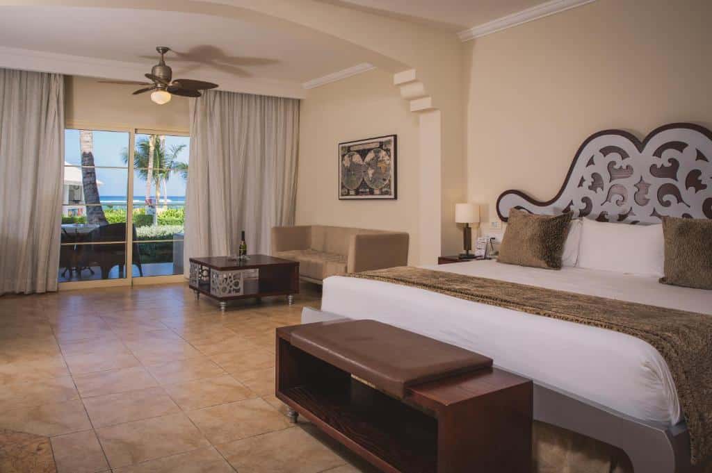 Quarto do Majestic Colonial Punta Cana, um dos hotéis românticos em Punta Cana. Uma cama de casal tem mesinhas de cabeceira e abajures dos dois lados, e um banquinho aos pés. Ao fundo do quarto, uma área de estar com mesinha de centro e sofá tem uma janela de vidro com vista para a varanda e para a praia.