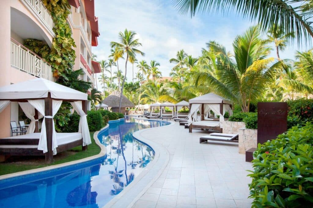 Piscina do Majestic Elegance Punta Cana rodeada por espreguiçadeiras e camas balinesas, que são estruturas relaxantes com colchão e cortinas nas laterais. Várias palmeiras ficam ao redor, e o prédio do hotel está no canto esquerdo.