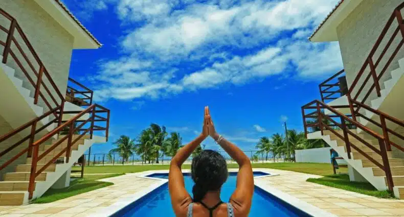 Pessoa de frente para uma piscina com as mãos levantadas em uma pose de ioga entre dois edifícios sob um céu azul brilhante com nuvens, no Manga Verde Beach Residence, para representar pousadas em Itamaracá (PE).