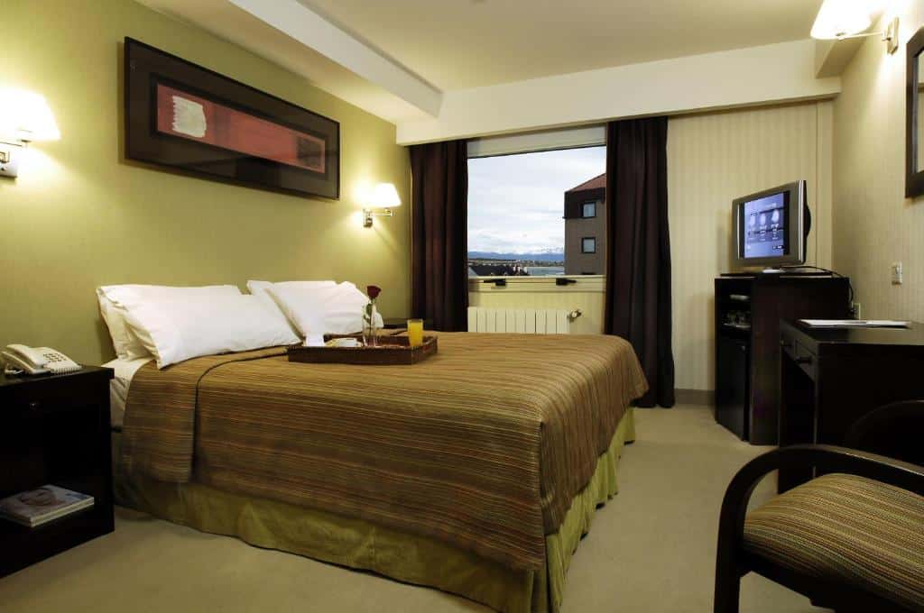 Quarto do MIL810 Ushuaia Hotel. Uma cama de casal do lado esquerdo, com um abajur de cada lado, de frente duas cômodas, uma televisão e uma cadeira. No fundo do quarto, a janela. Foto para ilustrar post sobre hotéis em Ushuaia.