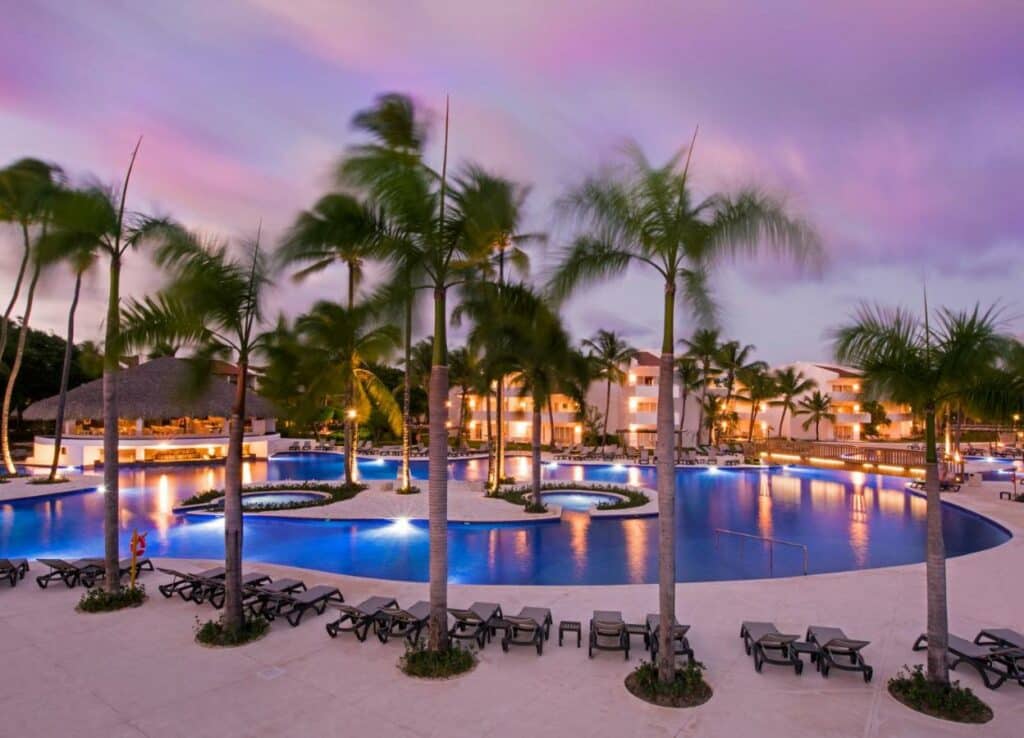 Piscina do Occidental Punta Cana ao pôr do sol. O local é rodeado por espreguiçadeiras e palmeiras, e ao fundo ficam os prédios do resort.