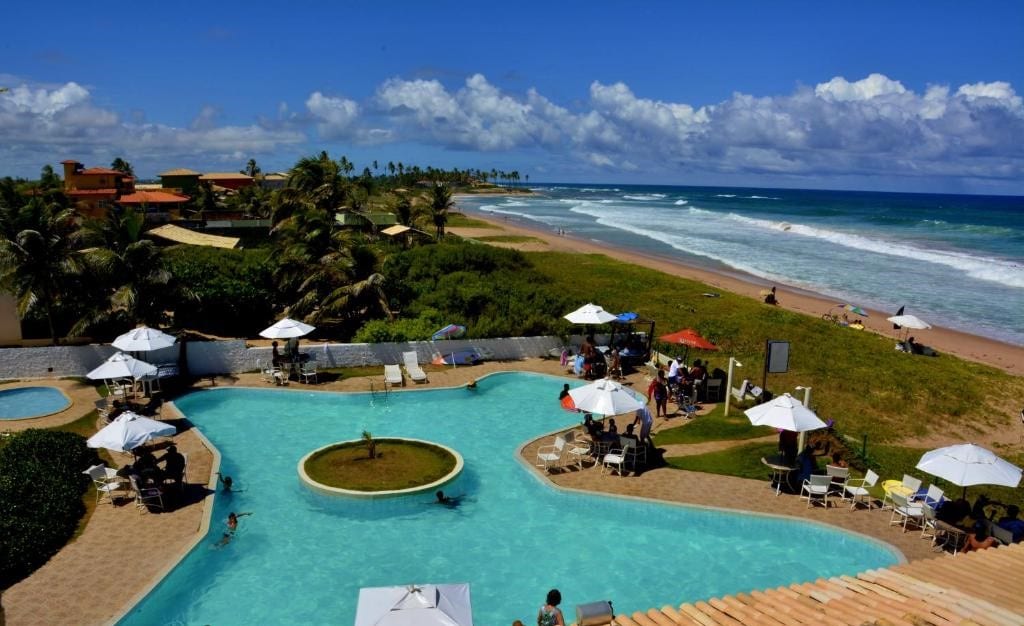 Piscina do OYO Hotel Arembepe Beach Hotel, Camacari. Uma piscina no meio, no centro dela uma ilha com grama, no lado esquerdo uma mini piscina, no lado direito a praia. Ao redor da piscina guarda-sóis, cadeiras e pessoas.