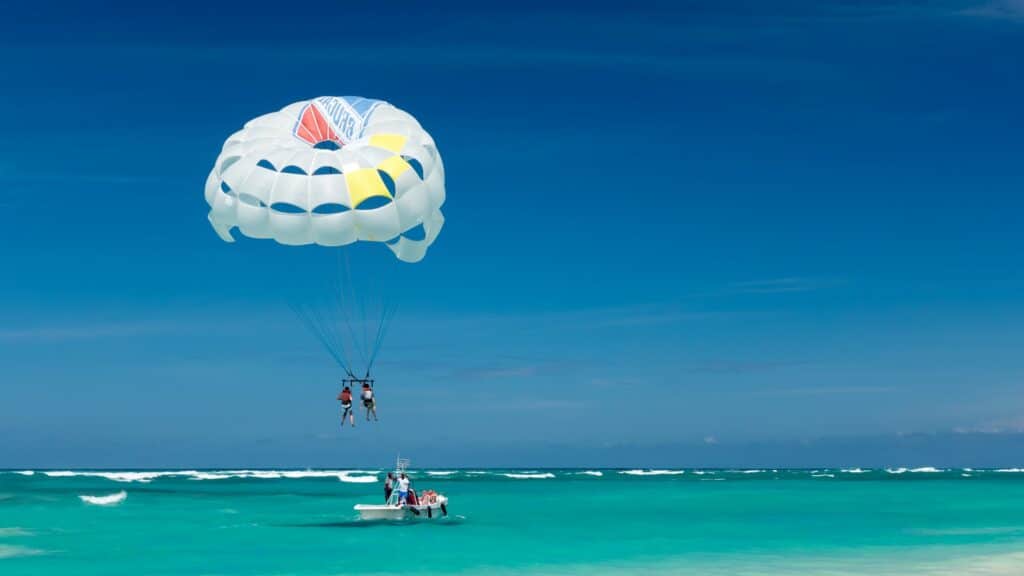 Duas pessoas voando com parasailing acima do mar em uma praia de Punta Cana. Embaixo deles há um barquinho com outras pessoas dando apoio ao esporte. O céu e o mar são azuis e se encontram no horizonte. - Foto: Joe deSousa via Unsplash