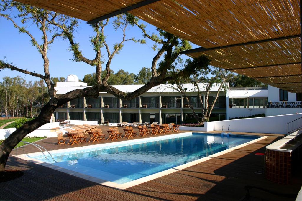 Piscina do Alentejo Star Hotel - Sao Domingos - Mertola. A piscina é retangular, no lado esquerdo há uma árvore, no lado direito é possível ver um pouco do bar e ao fundo estão cadeiras, mesas e espreguiçadeiras.