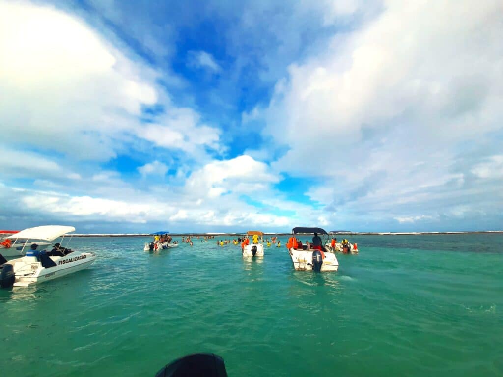 Piscina natural Ponta do Mangue em Maragogi, sendo o local com mar esverdeado e transparente, com alguns barcos navegando e pessoas nadando. Representa o que fazer em Maceió