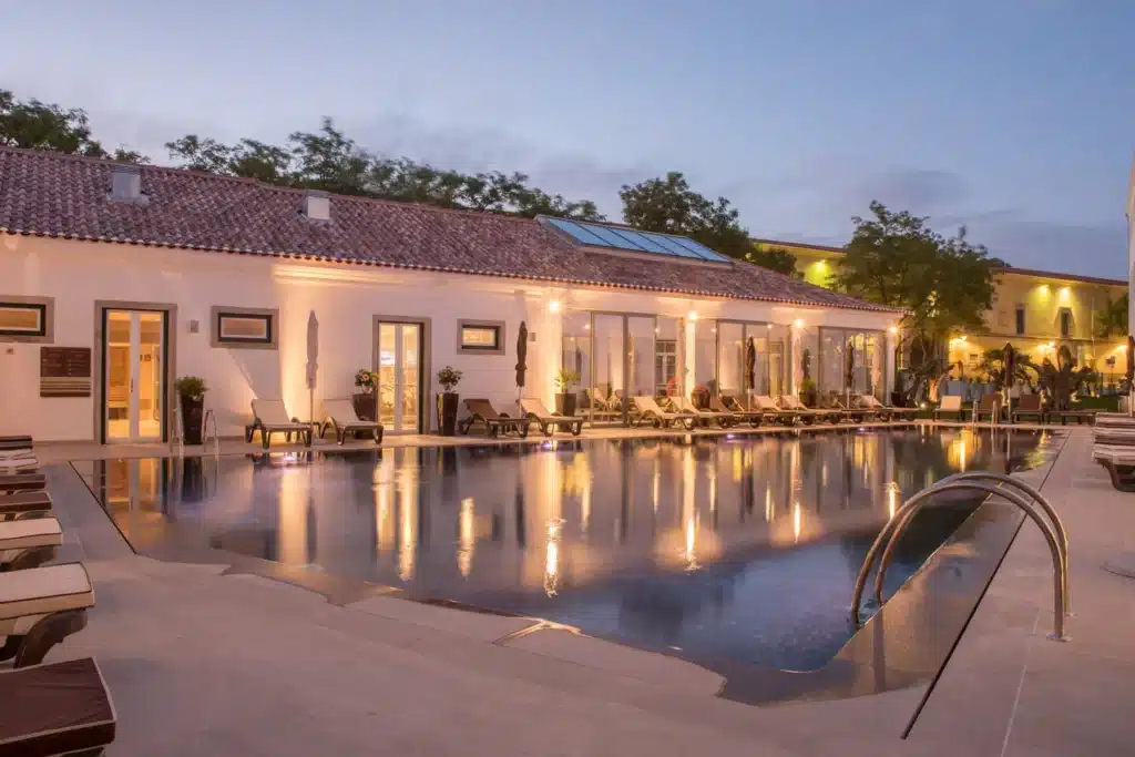 Área externa do hotel Vila Gale Collection Elvas. A frente há uma piscina com espreguiçadeiras e ao fundo há uma área fechada com a piscina coberta.