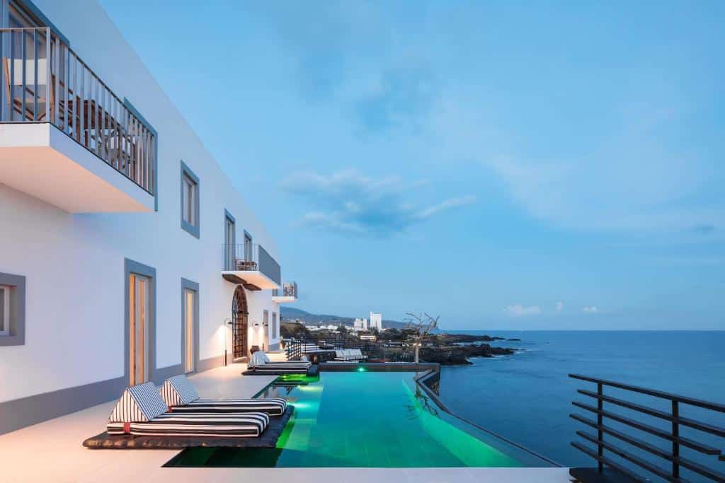 Piscina ao ar livre do White Exclusive Suites & Villas com a hospedagem do lado esquerdo da imagem com cadeiras a frente na borda da piscina que oferece vista para o mar.