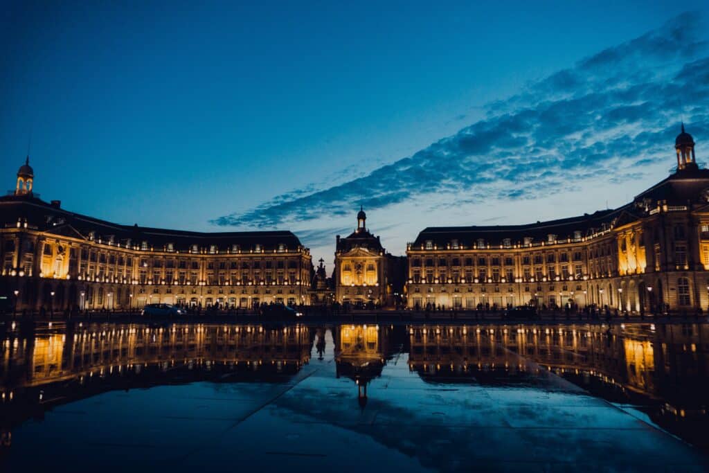 Place de la Bourse em Bordeaux. É uma construção grande e clássica, com vários detalhes e iluminada por luz amarela. Logo em frente está o espelho d'água e, como a foto foi tirada de noite, ele está refletindo a construção e o céu.