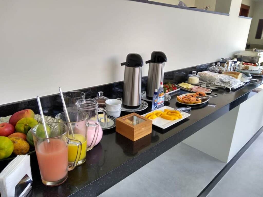 Café da manhã da Pousada Aromas da Pedra. Em um balcão estão jarras de suco, frutas, garrafas de café, frio, pães e outras comidas.