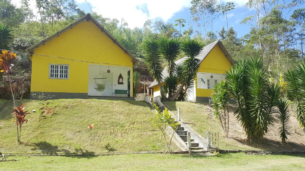 Foto externa dos dois chalés em Belo Canto Chalés. As casas são amarelas e possuem um varanda com banco e cadeira de balanço. Ambas acomodações estão localizadas no topo de um morro, com acesso por uma escadaria de cimento. Ao redor delas há bastante árvores e vegetação.