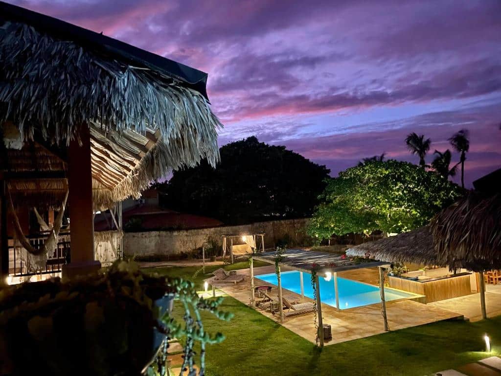 Vista de um dos quartos da Pousada Casa Caju Bangalows para a área ao ar livre com uma piscina ao centro, gramado verde e algumas árvores ao anoitecer, ilustrando post Pousadas em Cumbuco.
