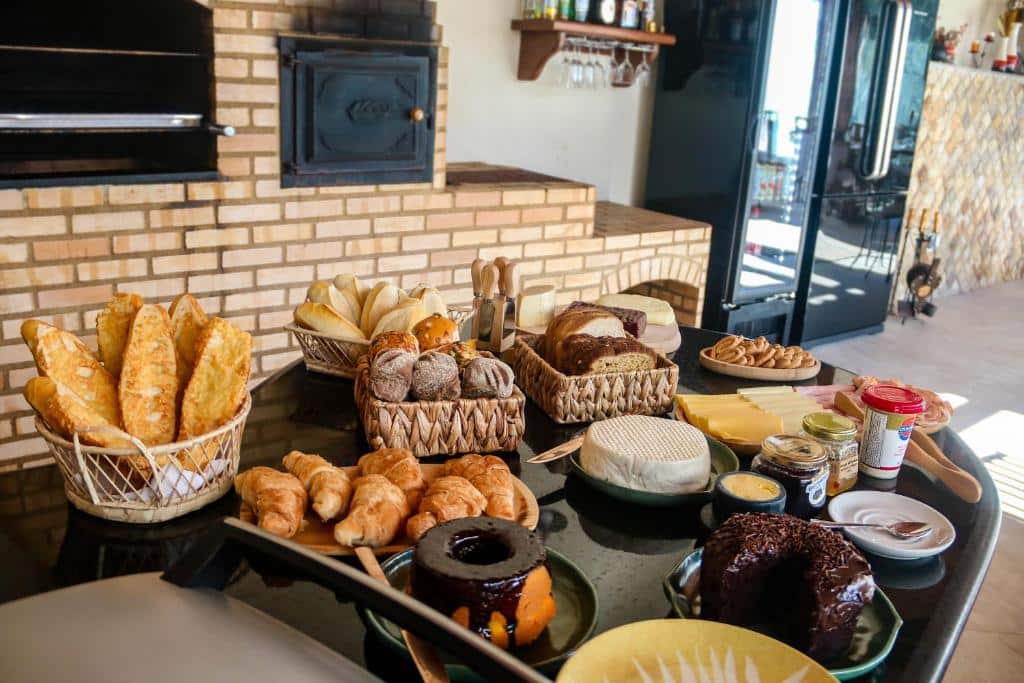 Café da manhã da Pousada dos Sonhos - MP. Uma mesa com pães, queijos, bolos e frios. Atrás uma churrasqueira e fogão a lenha. Do lado direito no fundo um freezer.