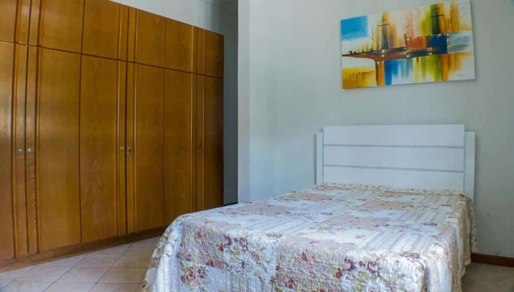 Foto de um quarto na Pousada Il Conventino, para ilustrar o post de pousadas em Santa Teresa (ES). Uma cama de casal com cabeceira branca de madeira está encostada em uma parede, e um quadro colorido está fixado acima dela. No lado esquerdo da foto, também encostado na parede, há um grande guarda-roupa de madeira. Há um bom espaçamento entre os móveis.
