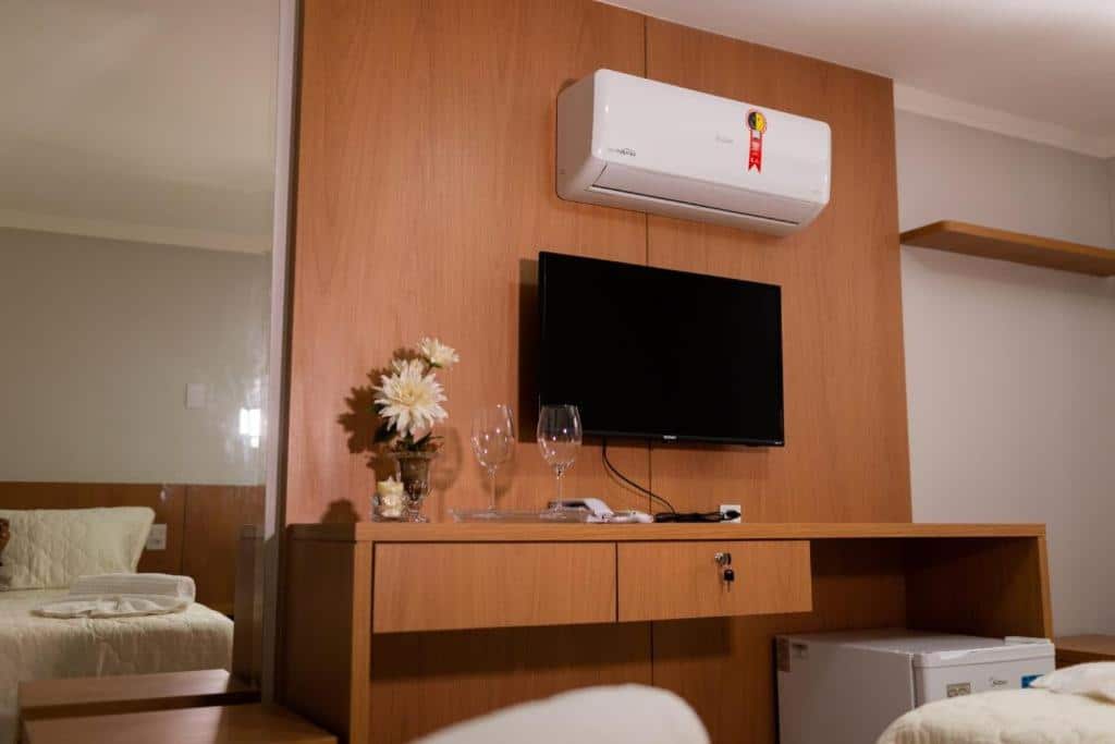 Outra visualização da suíte da Pousada La Sofia. É possível ver o painel de madeira que suporta uma televisão, ar condicionado, mesa com gavetas, objetos de decoração como taças e vaso com flores e um frigobar embaixo.